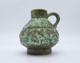 May include: A small, textured ceramic pitcher with a handle. The pitcher has a bulbous body and a short neck. The exterior is a mottled mix of green, brown, and gold, giving it a rustic appearance. The handle is a simple loop.