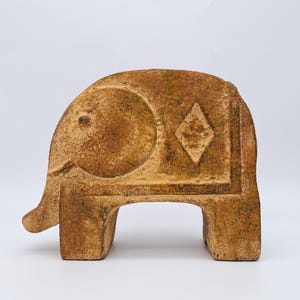 May include: A brown, textured, stone-like elephant figurine with a flat, blocky body. The side features a circular ear detail and a diamond-shaped carving. The surface has a weathered, mottled appearance with variations in brown and tan tones.