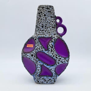 May include: A vintage ceramic vase with a textured black and white speckled finish. The vase features large, irregular purple shapes and a purple handle. The label on the vase reads "roth Keramik".