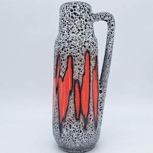 May include: A tall ceramic vase with a handle. The vase features a speckled black and white pattern with vertical red and black accents. The handle is integrated into the design, and the vase has a wide base.
