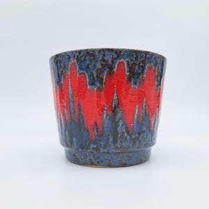 May include: A ceramic plant pot with a blue base and red, vertical, flame-like patterns. The pot has a textured surface and a slightly flared shape. The interior is a dark brown colour.