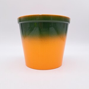 Peut inclure: Pot de plante en céramique avec un dégradé de couleurs. Le bord supérieur est vert foncé, passant à un corps orange moucheté. Le pot est cylindrique et posé sur un fond blanc.