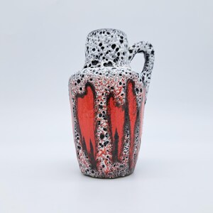 May include: A ceramic vase with a handle, featuring a textured surface. The top is white with black speckles, transitioning to a vibrant red base with black accents. The vase has a unique, artistic design.