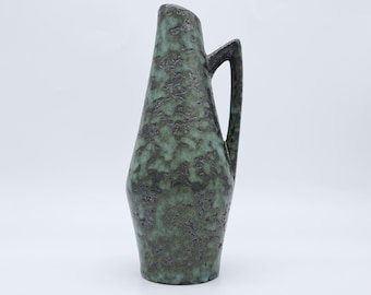 May include: A tall ceramic pitcher with a tapered silhouette and a single handle. The surface features a mottled, textured glaze in shades of dark charcoal gray and muted teal green, creating a layered, uneven appearance across the body.