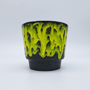 May include: A ceramic cup featuring a dark charcoal base with a vibrant, neon yellow glaze dripping from the rim down the sides. The vessel has a wide, open top that tapers towards a narrower, solid-coloured base, creating a distinct silhouette.