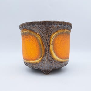 Peut inclure: Un pot en céramique au design unique. Il présente un centre orange vif avec une bordure texturée marron foncé et des éléments décoratifs. Le pot a une forme arrondie avec une base légèrement pointue.