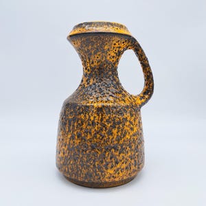 May include: A textured, orange and black ceramic pitcher with a handle. The pitcher has a wide base that tapers to a narrow neck, then flares out at the top. The handle is a curved loop. The surface has a speckled, rough texture.