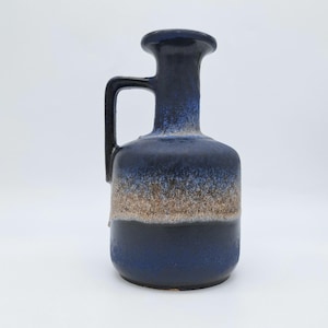Op de afbeelding: Een keramische vaas met een donkerblauwe glazuur, een handvat en een brede basis. De vaas heeft een cilindrische hals en een afgeronde bovenkant. Het middengedeelte gaat over in een bruine tint. Een decoratief stuk voor de woondecoratie.