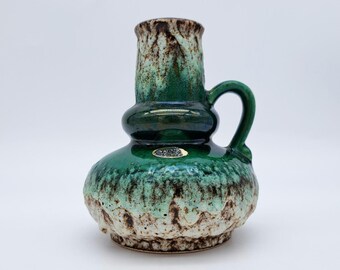 May include: A green and brown ceramic vase with a handle. The vase has a bulbous base and a cylindrical neck. The glaze has a mottled appearance, with shades of green and brown. A small sticker is visible on the front.