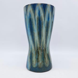 Könnte beinhalten: Eine hohe Keramikvase mit ausgestelltem Rand und schmaler Taille. Die Vase hat eine blau-grüne Glasur mit vertikalen, wellenförmigen Mustern. Der Sockel ist in einem dunkleren Blauton gehalten.