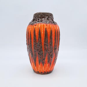 May include: A vintage ceramic vase with a textured surface. The vase has a vibrant orange and dark brown colour scheme with a geometric pattern. The top of the vase has a dark brown, porous texture.