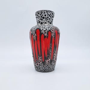 May include: A decorative ceramic vase with a textured surface. The vase features a striking design with vertical red stripes against a black and white speckled background. The vase has a cylindrical body and a flared neck.