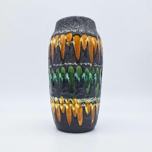 May include: A tall, textured black ceramic vase with bands of orange and green drip glaze. The vase has a cylindrical shape with a slightly wider base and a narrower neck. The glaze creates a unique, artistic design.