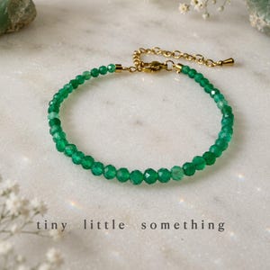 Op de afbeelding: Een delicate groene kralenarmband met een goudkleurige ketting en sluiting. De armband is gemaakt van kleine, facetgeslepen groene kralen. De tekst "tiny little something" is in een lichte kleur onderaan de afbeelding gedrukt. De armband ligt op een wit marmeren oppervlak.