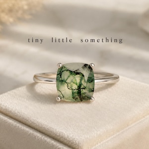 May include: A silver ring featuring a square, moss-green gemstone. The ring is displayed on a cream velvet surface. The text "tiny little something" is above the ring.