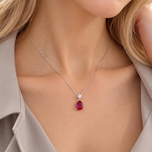 May include: A silver-toned chain necklace featuring a teardrop-shaped red gemstone pendant suspended below a small, clear, round-cut stone. The text "tiny little something" is printed in small, black, lowercase letters at the bottom center.