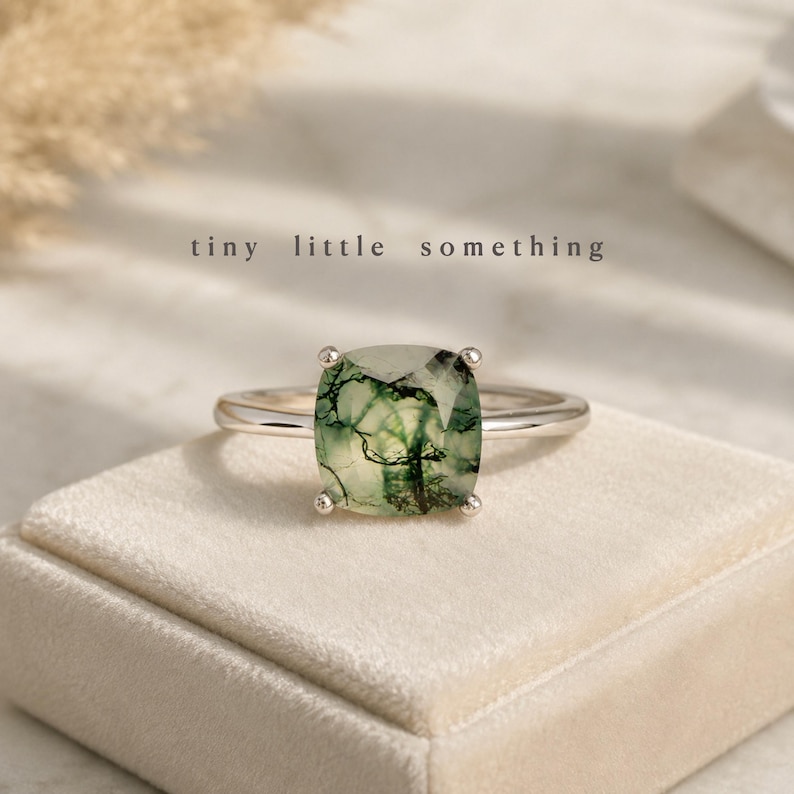 Puede incluir: Un anillo de plata con una gema verde musgo cuadrada. El anillo est&aacute; sobre una superficie de terciopelo crema. El texto "tiny little something" est&aacute; encima del anillo.