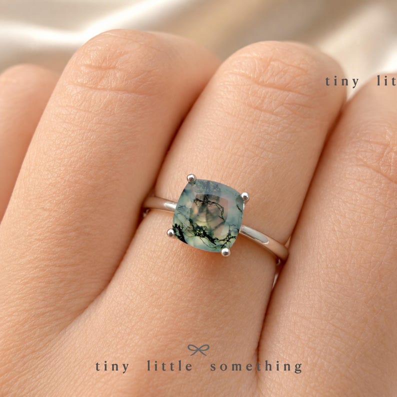 Puede incluir: Un anillo de plata con una piedra preciosa de &aacute;gata musgosa de corte cuadrado. La piedra muestra inclusiones verdes y negras, engastada en una delicada banda de plata. El texto "tiny lit" y "tiny little something" est&aacute; impreso en la imagen.
