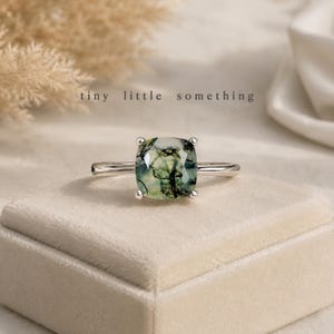 Puede incluir: Anillo de plata con una gema de &aacute;gata musgosa de corte cuadrado, con inclusiones verdes y negras. El anillo se muestra sobre una superficie de terciopelo beige claro. El texto "tiny little something" est&aacute; presente.