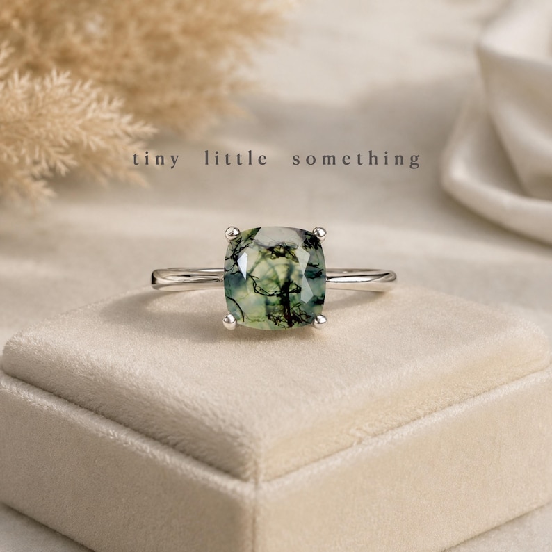 Puede incluir: Anillo de plata con una gema de &aacute;gata musgosa de corte cuadrado, con inclusiones verdes y negras. El anillo se muestra sobre una superficie de terciopelo beige claro. El texto "tiny little something" est&aacute; presente.