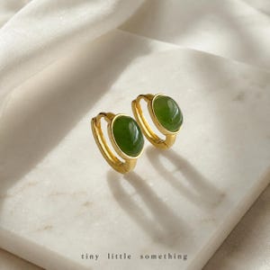 May include: A pair of gold hoop earrings featuring smooth, oval-shaped green jade stones set in the centre. The earrings have a polished metallic finish and a hinged closure mechanism. The text "tiny little something" is printed at the bottom.