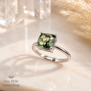 Puede incluir: Un anillo de plata con una piedra preciosa verde cuadrada. La piedra tiene un patr&oacute;n &uacute;nico similar al musgo. El anillo est&aacute; sobre una superficie blanca con el texto "tiny little something" en la parte inferior.