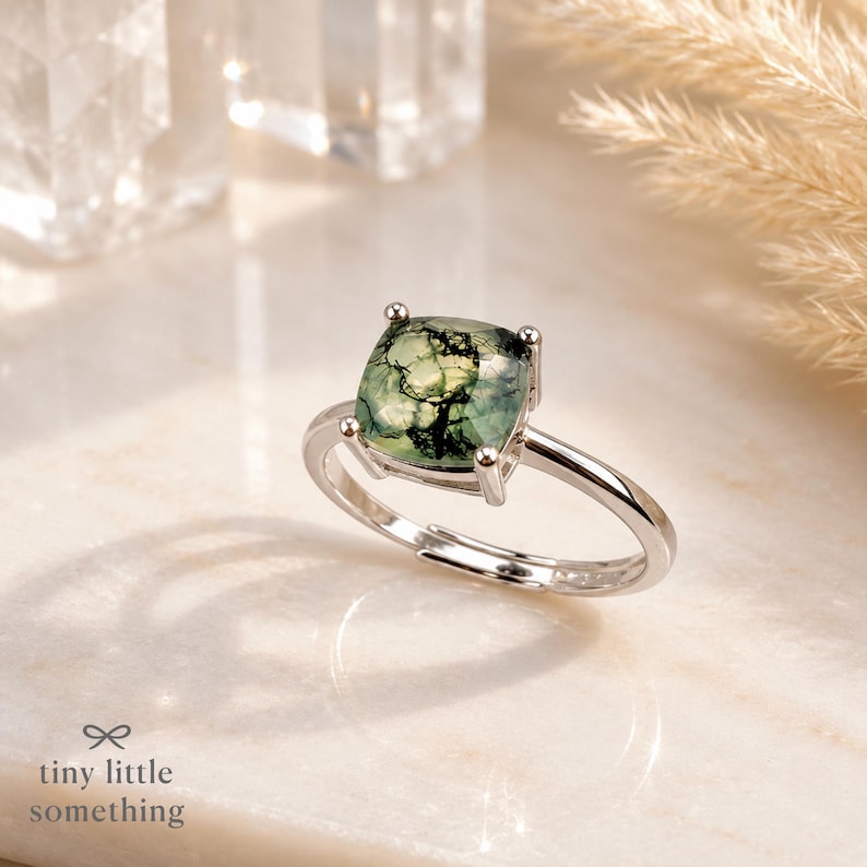 Puede incluir: Un anillo de plata con una piedra preciosa verde cuadrada. La piedra tiene un patr&oacute;n &uacute;nico similar al musgo. El anillo est&aacute; sobre una superficie blanca con el texto "tiny little something" en la parte inferior.