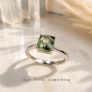 Puede incluir: Un anillo de plata con una piedra cuadrada de &aacute;gata musgosa. La piedra es una mezcla de verde y blanco con vetas oscuras. El anillo est&aacute; sobre una superficie blanca, con la frase "tiny little something" en la parte inferior.