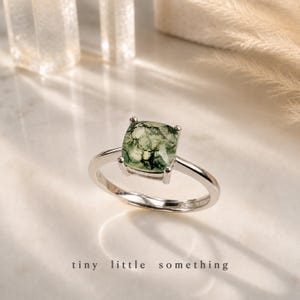 Puede incluir: Un anillo de plata con una piedra de &aacute;gata musgosa verde cuadrada. La piedra tiene un patr&oacute;n natural &uacute;nico. El anillo est&aacute; sobre una superficie blanca con las palabras "tiny little something" en la parte inferior.