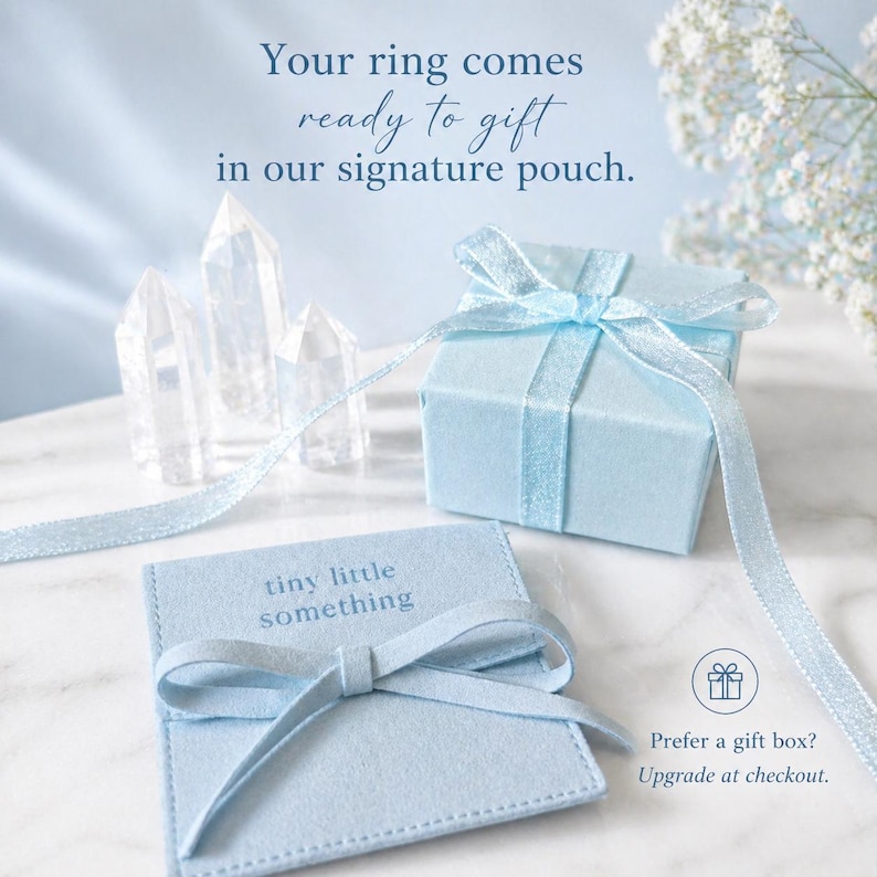 Puede incluir: Una caja de regalo azul claro atada con una cinta a juego, junto a una peque&ntilde;a bolsa con el texto "tiny little something". La imagen tambi&eacute;n incluye formaciones de cristal transparente y el texto "Your ring comes ready to gift in our signature pouch."