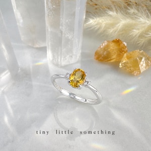 May include: A silver ring featuring an oval yellow gemstone and two smaller white gemstones. The ring is set against a soft background with the text "tiny little something".