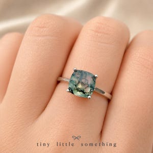 Puede incluir: Anillo de plata con una gema de &aacute;gata musgosa de corte cuadrado. La piedra muestra tonos verdes, azules y blancos con patrones intrincados. El texto "tiny little something" est&aacute; en la parte inferior.