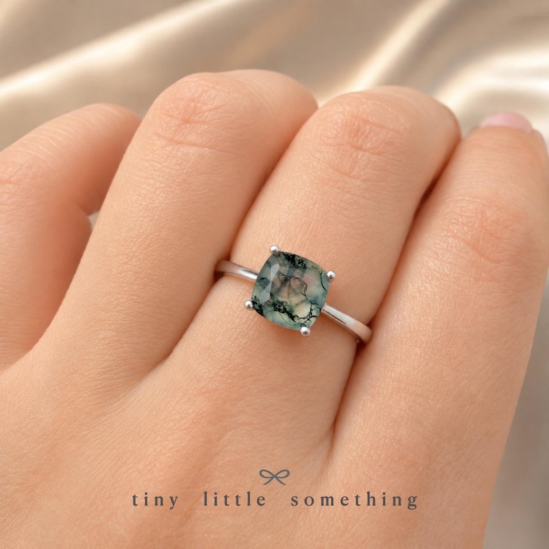 Puede incluir: Un anillo de plata con una piedra de &aacute;gata musgosa de corte cuadrado. La piedra muestra patrones verdes y blancos. El anillo se muestra en una mano, con el texto "tiny little something" en la parte inferior.
