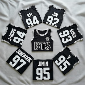 May include: Black basketball jerseys with white trim and lettering. The jerseys feature names and numbers, including "RM 94", "JIN 92", "J-HOPE 94", "SUGA 93", "JUNGKOOK 97", "JIMIN 95", and "V 95". A central jersey displays "BTS".
