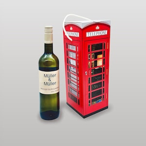 Könnte beinhalten: Eine grüne Weinflasche mit weißem Etikett neben einer roten Telefonzellen-Geschenkbox. Auf der Geschenkbox steht das Wort „TELEPHONE“. Die Weinflasche hat einen goldfarbenen Verschluss.