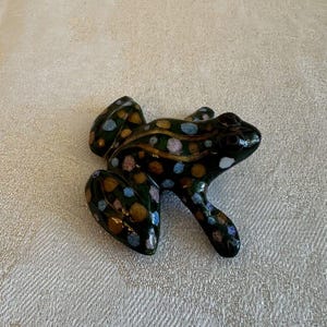 May include: A ceramic frog figurine with a black base and colorful spots in shades of blue, pink, and gold.