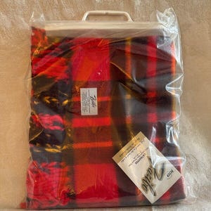 May include: A red, black, and yellow plaid throw blanket in a plastic bag with a white handle. The bag has a label that says "Carriere 100% Acrylic" and "Zella"