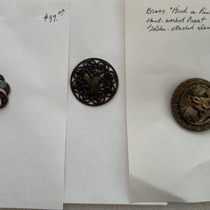 May include: Three antique brass buttons with intricate designs. The first button is a swirl pattern with red, blue, and white enamel. The second button is a bird in a tree design. The third button is a bird in a tree design with a more detailed, textured finish. The buttons are priced at $39.00 and $29.00.