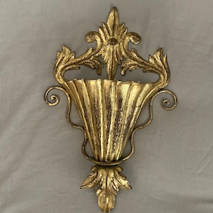 May include: Ornate gold-colored wall sconce with a fluted, vase-like design. The sconce features decorative leaf and scroll accents, and a textured finish. This vintage-style piece is designed for wall mounting and adds a touch of elegance.