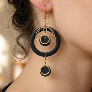 Può includere: Un orecchino pendente con un grande anello nero opaco sospeso a un gancio dorato. All'interno dell'anello grande ci sono due anelli dorati più piccoli e un piccolo cerchio nero. Un secondo piccolo cerchio nero pende dalla parte inferiore.