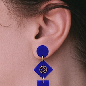 Può includere: Orecchini blu geometrici con un cerchio, un rombo con un cerchio dorato e un quadrato. Gli orecchini sono collegati con metallo color oro. Gli orecchini sono mostrati su un orecchio.