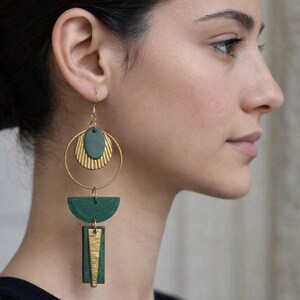 Può includere: Orecchini pendenti composti da un cerchio in metallo dorato, un disco ovale verde scuro con un semicerchio dorato testurizzato, un elemento semicircolare verde scuro e un pendente rettangolare verde scuro con un inserto triangolare dorato.