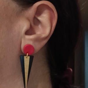 Può includere: Orecchini geometrici con un cerchio rosso, un triangolo nero con una striscia dorata e un filo di collegamento dorato. Gli orecchini sono mostrati vicino a un orecchio.