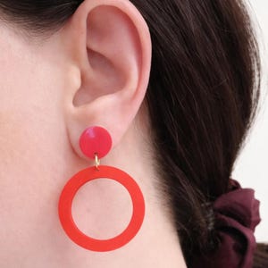 Può includere: Orecchini a cerchio rossi con un piccolo bottone rosso. Gli orecchini sono di un rosso brillante e pendono da un perno dorato. Gli orecchini sono mostrati su un orecchio.