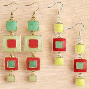 Può includere: Due paia di orecchini pendenti con ciondoli quadrati sovrapposti. La coppia a sinistra ha quadrati più grandi in verde, rosso e crema. La coppia a destra ha quadrati più piccoli in giallo, rosso e verde. Tutti i pezzi sono uniti da maglie dorate.