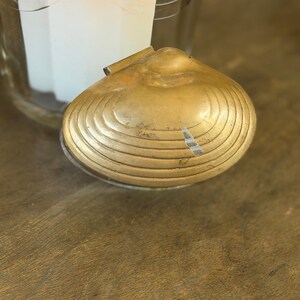 May include: A vintage brass clam shell-shaped trinket box with a hinged lid. The box has concentric circular ridges and a tarnished finish, suggesting age. The background includes a glass container with a white interior.