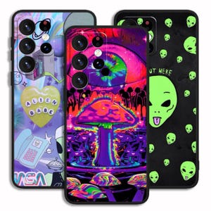 May include: Three different phone cases with colorful designs. The first case has a pink and blue alien theme with the text "Alien Babe". The second case has a psychedelic mushroom design with a rainbow eye. The third case has a black background with green alien faces and the text "Out Here".
