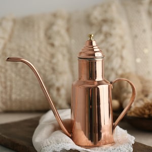 Puede incluir: Un dispensador de aceite de cobre con una tapa de latón y un pico largo. El dispensador está sobre una tabla de madera con un paño blanco.