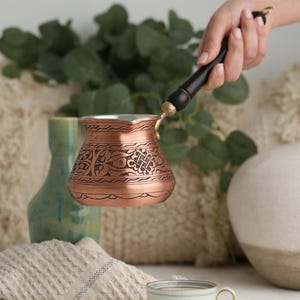 Puede incluir: Una cafetera de cobre con un diseño adornado y un mango de madera. La cafetera se está vertiendo en una taza de café blanca con un borde dorado.