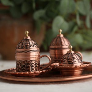 May include: A set of three copper Turkish coffee cups and saucers with intricate designs. The cups have domed lids and the saucers have a scalloped edge.